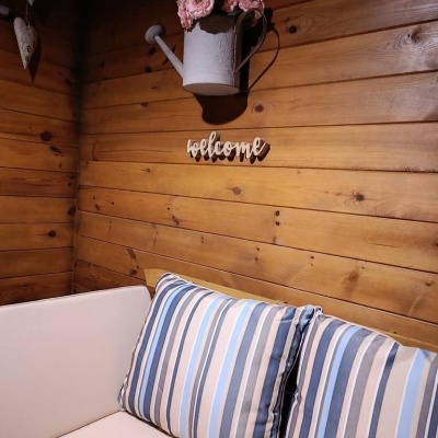 Sofá branco com almofadas às riscas azuis junto a parede de madeira com regador com flores e placa 'welcome'