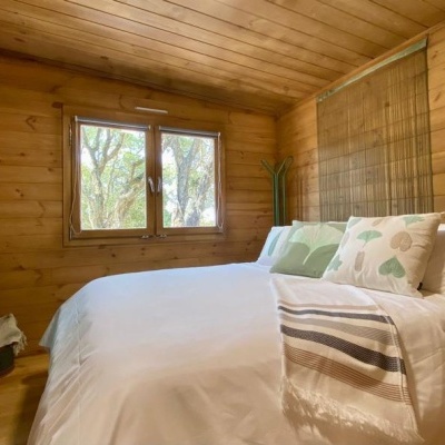 Quarto com cama de madeira, roupa branca e almofadas verdes, paredes e teto de madeira, janela com vista para árvores