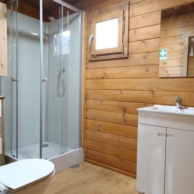 Casa de banho com madeira clara, cabine de duche em vidro, sanita e móvel de lavatório branco.