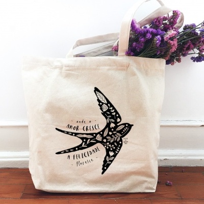 Saco de pano bege com ilustração preta de andorinha e texto, flores roxas e rosas dentro