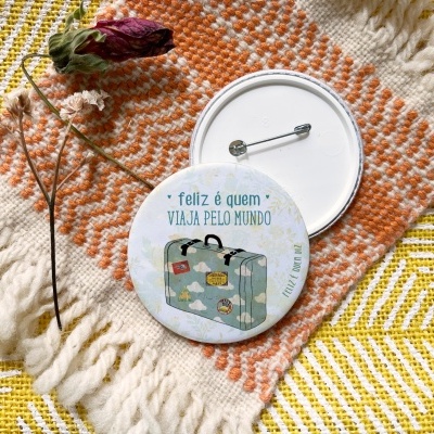 Botão branco com desenho de mala azul e texto, em tecido têxtil laranja e amarelo com franjas.