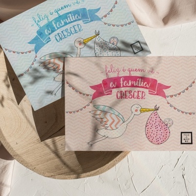 Cartões de felicitações azuis e rosa com ilustração de cegonha e texto sobre mesa branca e tábua de madeira