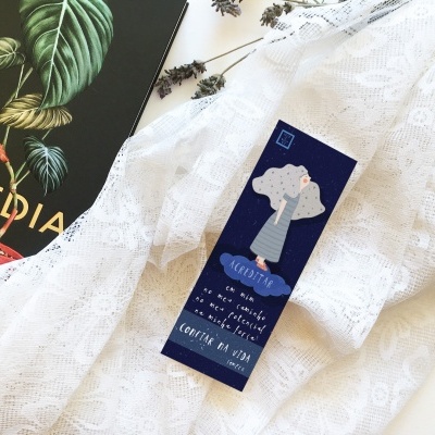 Marcador de páginas azul com ilustração de árvore sobre tecido branco rendado, ao lado livro preto com a palavra TOPEDIA.