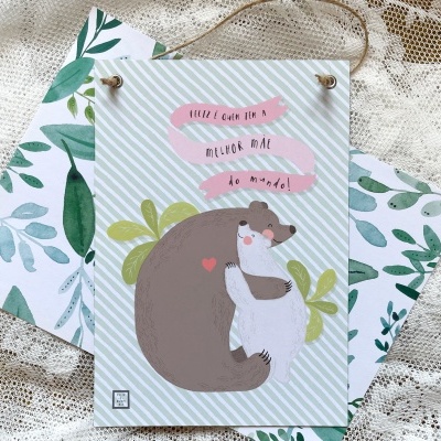 Placa decorativa com ilustração de urso e texto em inglês pendurada com corda sobre fundo de tecido e caixa com padrão de folhas verdes