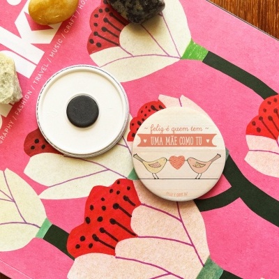 Espelho de bolsa redondo com capa decorativa rosa e bege, sobre revista cor-de-rosa com flores e três pedras