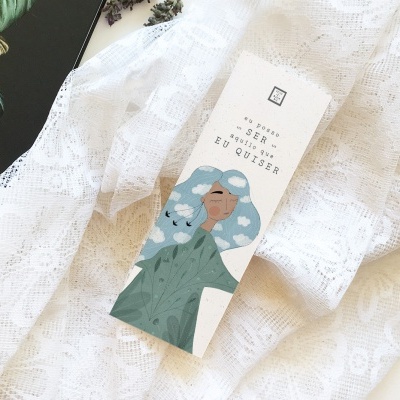 Embalagem branca com ilustração colorida e texto, sobre tecido rendado branco e livro com folhagens