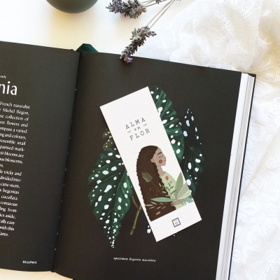 Marcapáginas com ilustração de mulher e folhas verdes em livro aberto ao lado de tecido branco e lavanda