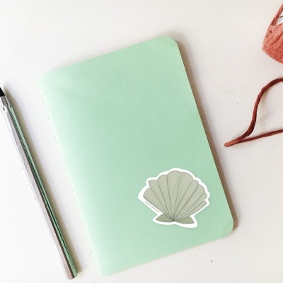 Caderno verde com autocolante concha, caneta prateada, fio castanho e pássaro decorativo cor de coral numa superfície branca.