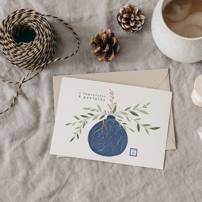 Cartão ilustrado com vaso azul e plantas, envelope, fio, pinhas e chávena de café sobre tecido cinzento