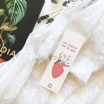 Marcador de livros com desenho de coração e texto, sobre tecido branco rendado, junto a livro preto com título TOPEDIA.