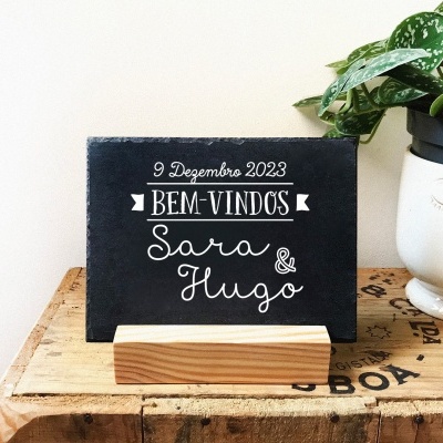 Placa preta com texto branco e vaso branco com plantas verdes sobre caixa de madeira rústica