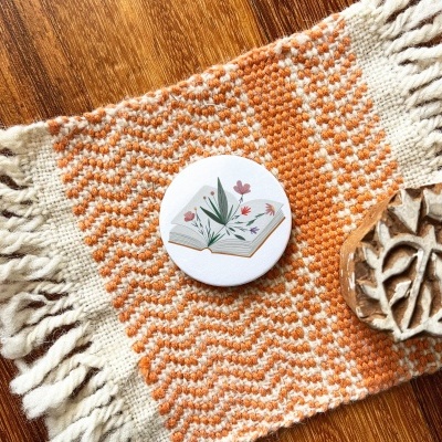 Botão redondo decorativo com ilustração floral sobre passadeira laranja e branca com franjas e peça esculpida em madeira