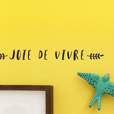 Parede amarela com o texto decorativo 'JOIE DE VIVRE' e um pássaro azul em relevo