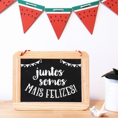 Lousa com mensagem em português, vaso com planta e guirlanda de melancias decorativa numa mesa de madeira