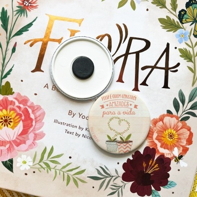 Livro infantil 'Flora' com capa florida e dois botões brancos sobre tecido amarelo e branco
