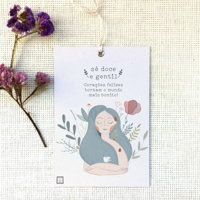 Etiqueta decorativa com ilustração de mulher e texto motivacional em português sobre fundo de tecido branco, com flores secas ao lado.