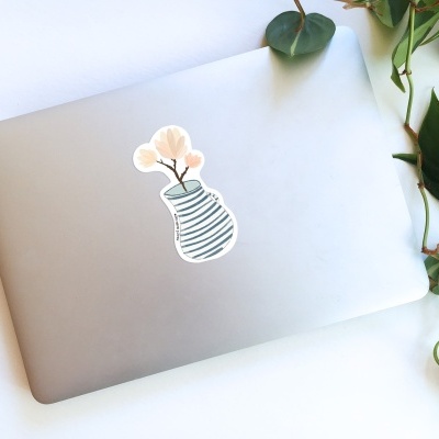 Computador portátil cinzento com autocolante de jarra e flores ao lado de plantas verdes