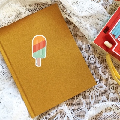 Caderno castanho com autocolante de gelado e câmara fotográfica vermelha em fundo de renda branca