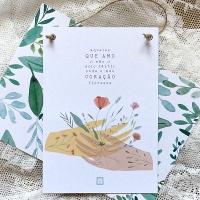 Cartão com mãos e flores, texto em português, sobre papel com folhas verdes