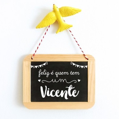 Placa decorativa com texto 'feliz é quem tem um Vicente' pendurada por cordão com pássaro amarelo