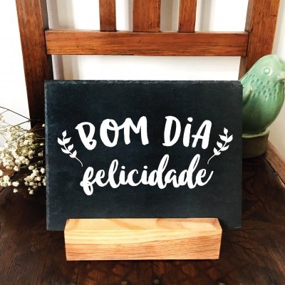 Placa decorativa preta com texto 'BOM DIA felicidade' numa cadeira de madeira com flores e pássaro