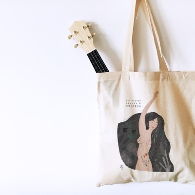 Saco de pano bege com estampa de mulher e texto, pendurado na parede com guitarra dentro.