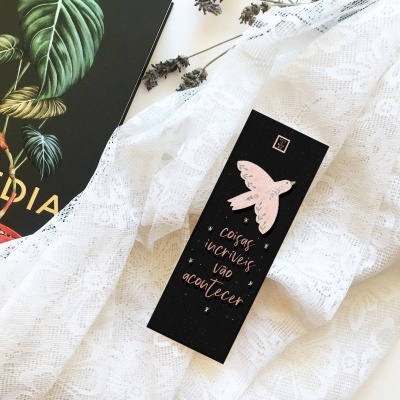 Marcador de livros preto com borboleta rosa e texto dourado sobre tecido branco rendado junto de livro preto com folhas