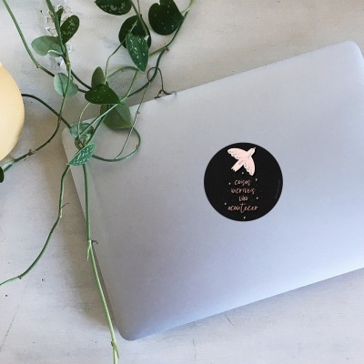 Computador portátil prateado com autocolante preto redondo com texto e estrela rosa, planta verde e jarro amarelo numa mesa branca.