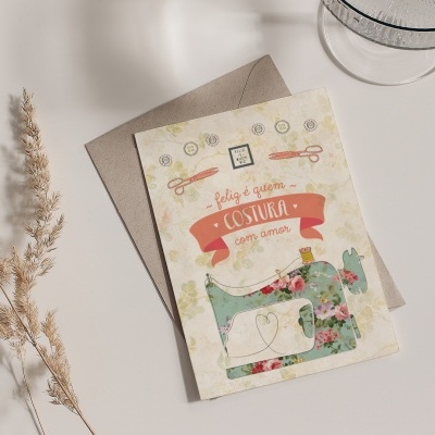 Cartão com design de máquina de costura e texto em português sobre mesa branca com planta seca e vidro