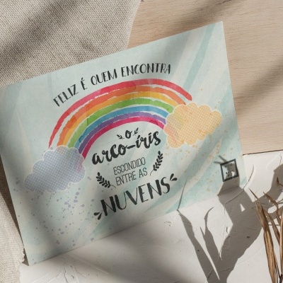 Postal colorido com arco-íris e frase em português sobre uma superfície branca, tecidos e ramo seco