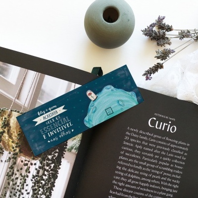 Livro aberto com texto em inglês, marcador azul com ilustração, vaso cinza e flores secas sobre superfície branca