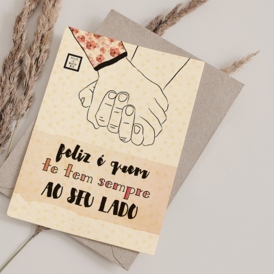 Cartão de felicitações com duas mãos dadas e texto em português, sobre envelope castanho e raminhos secos