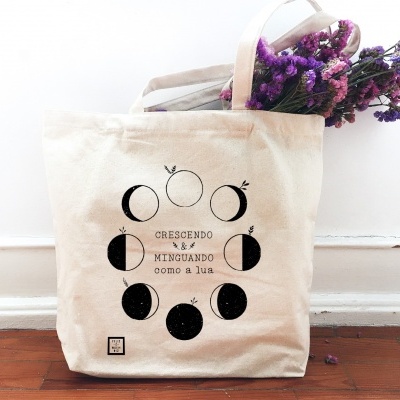 Saco de tecido cru estampado com fases da lua e texto, contendo flores roxas, sobre chão de madeira