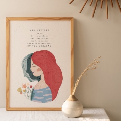 Pôster decorativo com duas mulheres desenhadas e texto em português numa moldura de madeira, sobre uma mesa clara com um vaso cerâmico ao lado.
