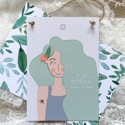 Cartão decorativo com ilustração de mulher e texto positivo, sobre tecido de renda e envelope com padrão de folhas verdes.