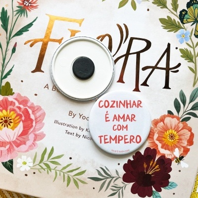 Livro com capa floral e borboleta, botão com frase em português e tampa metálica em tecido amarelo.