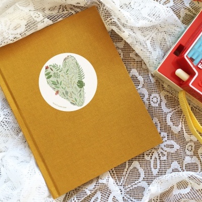 Caderno mostarda com ilustração de folhas sobre tecido rendado, ao lado de câmara fotográfica vermelha e branca