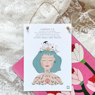 Cartaz decorativo pendurado com ilustração de mulher e texto motivacional, sobre tecido rendado e objeto rosa com flores.