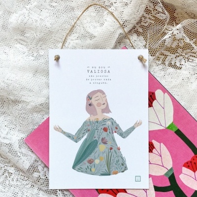 Cartaz pendurado com ilustração de mulher em vestido verde e texto, sobre tecido rendado branco e fundo rosa floral.