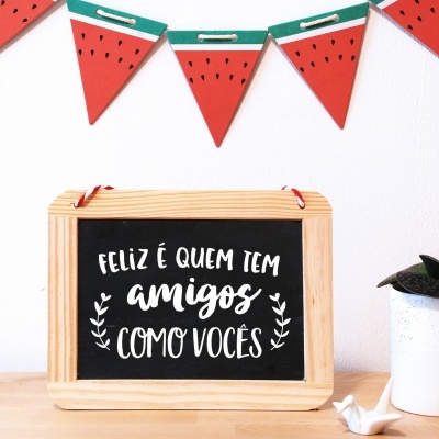 Quadro negro com mensagem em português e decoração de melancia na parede sobre mesa com vaso branco.