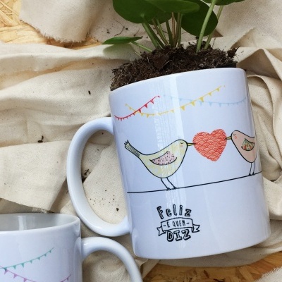 Caneca branca com desenho de passarinhos, coração vermelho, bandeirinhas coloridas e a palavra 'Feliz', com planta dentro.