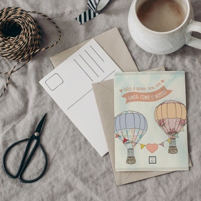 Cartão postal com balões coloridos, envelope, cartão vazio, fio em bobinas, tesoura e caneca numa mesa