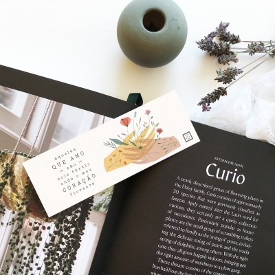 Livro aberto com texto e marcador de papel floral ao lado de vaso cinza e flores secas