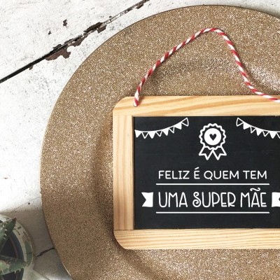 Quadro decorativo com frase em português sobre ser uma super mãe, moldura de madeira, pendurado com cordão, sobre tapete dourado e planta suculenta.