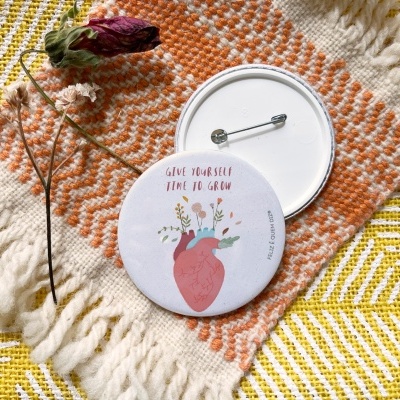 Dois botons redondos com alfinete, um ilustrado, sobre tecido com padrão e flor seca