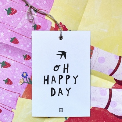 Etiqueta com texto 'OH HAPPY DAY' sobre tecidos coloridos com padrão de morangos e formas abstratas