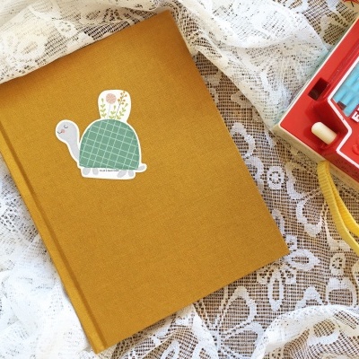 Caderno amarelo com autocolante de tartaruga e câmara Fisher-Price vermelha sobre renda branca