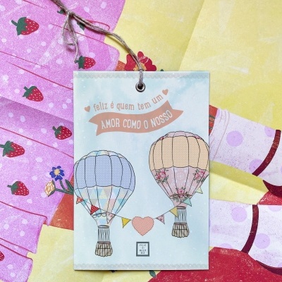 Cartão com balões de ar quente e frase, sobre tecidos coloridos com padrão de morangos.