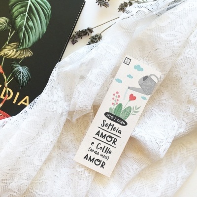 Marcador de livros decorativo com texto e ilustrações, sobre tecido branco rendado ao lado de livro preto