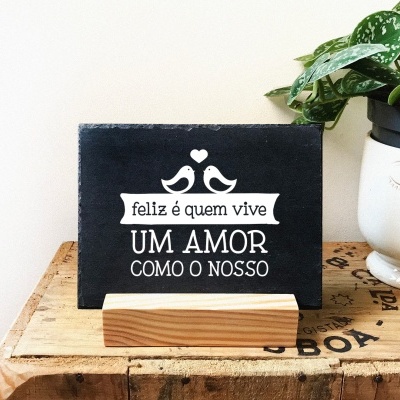 Quadro preto com texto branco e desenho de pássaros em suporte de madeira ao lado de vaso com planta